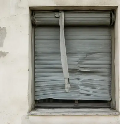 Volet roulant gris abîmé, lames déformées et sangle pendante